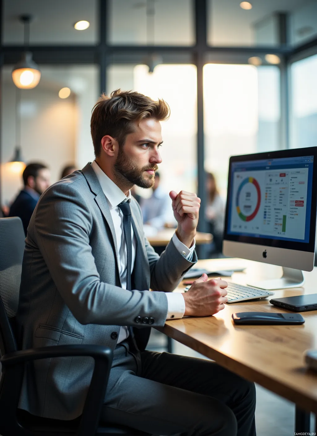 A man in a suit sits at a desk in an office, looking at a computer screen.