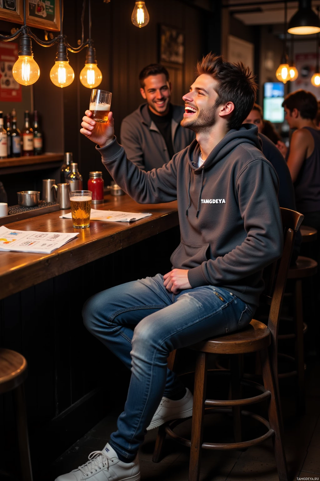 A man in a hoodie sits at a bar, holding a beer glass and smiling.
