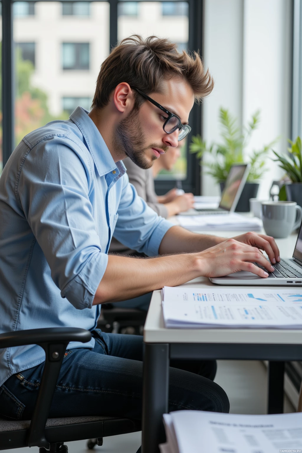 A man wearing glasses works on a laptop in an office setting.