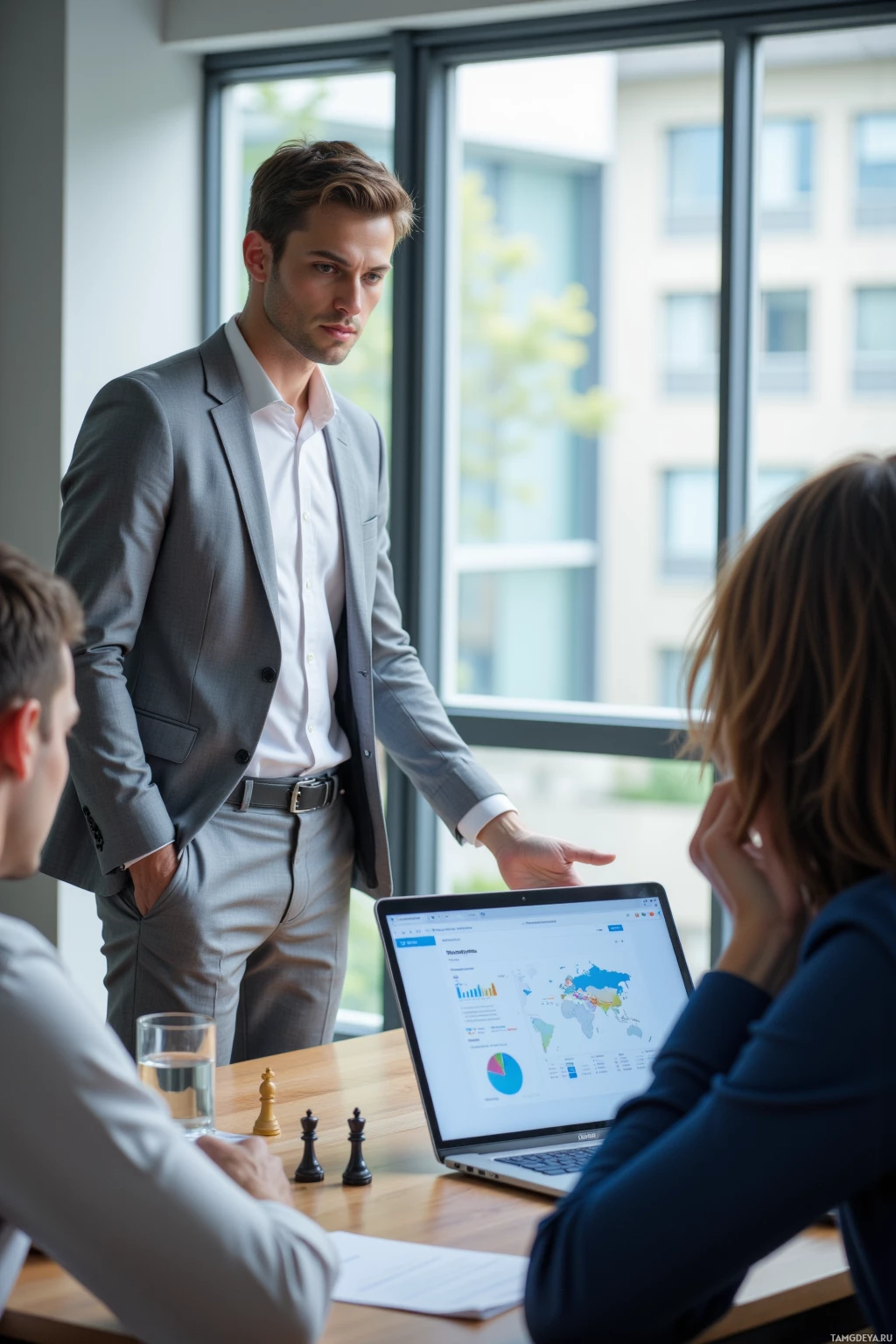 A man in a suit stands by a table with a laptop and chess pieces, discussing with two others.