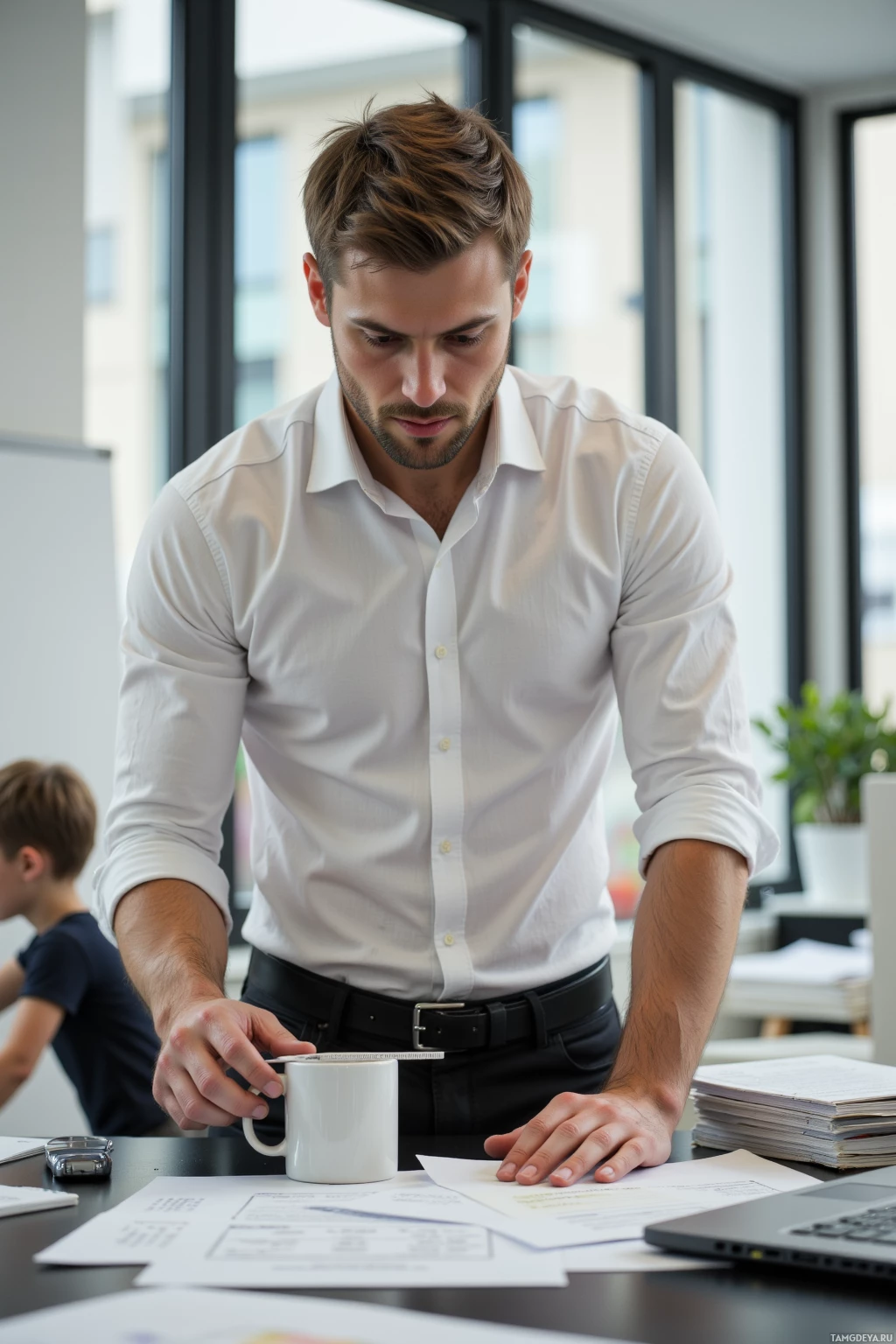 A man in a white shirt leans over a desk with papers and a laptop, appearing focused.