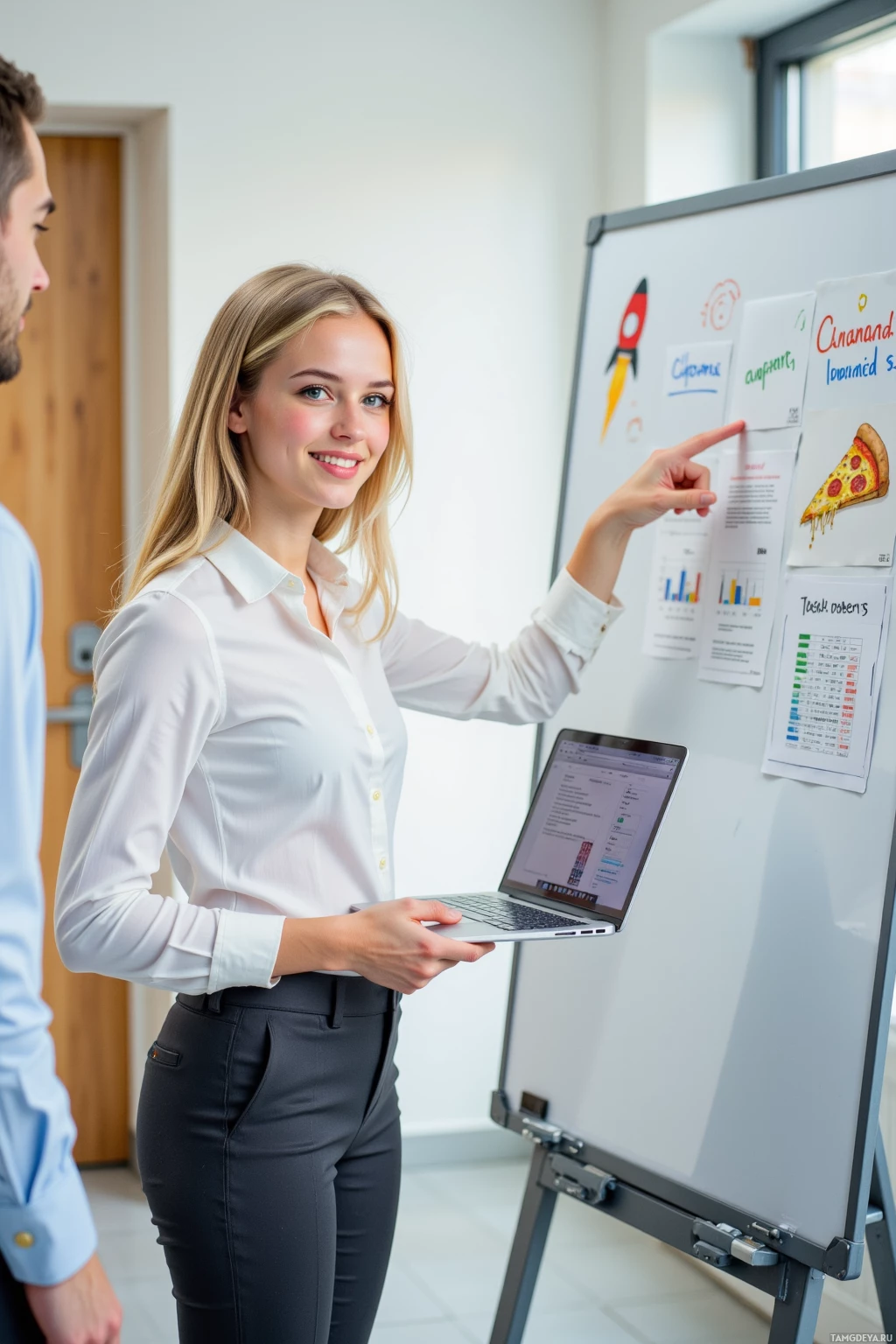 A woman in a white shirt and black pants stands in front of a whiteboard, pointing at it while holding a laptop.