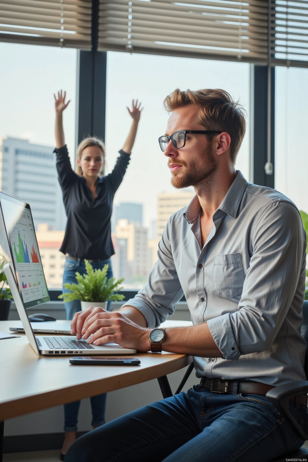 A man works on a laptop while a woman raises her hands in the background.